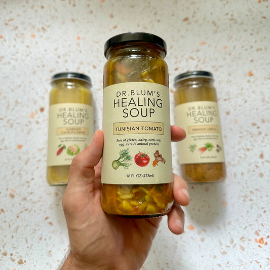 Hand holding a jar of Dr. Blum's Healing Soup with two other jars in the background on a speckled countertop.